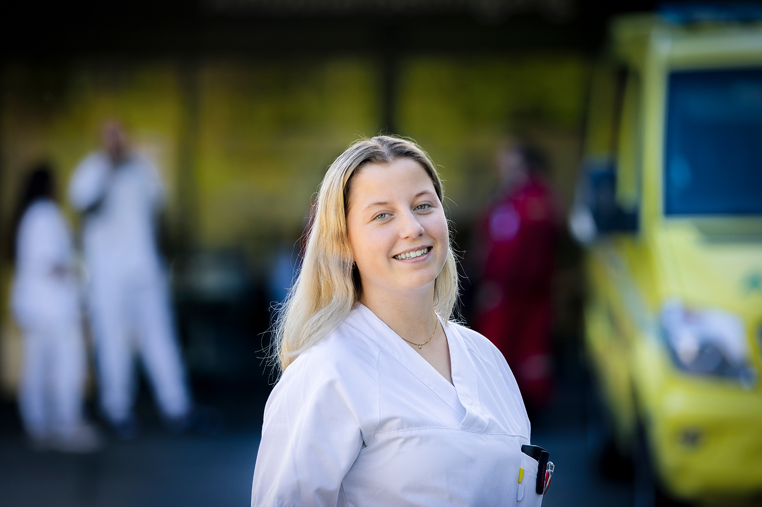 Helsearbeider i hvit uniform smiler foran en ambulanse.