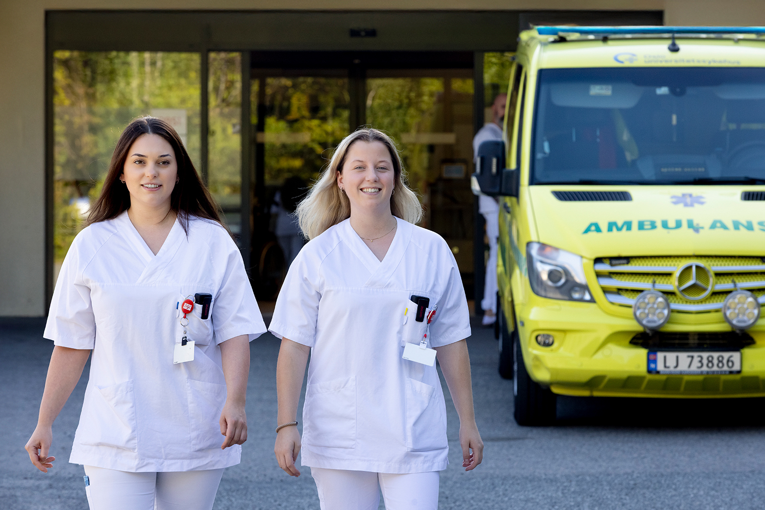 To helsearbeidere i hvite uniformer går ut av en bygning. En gul ambulanse står parkert bak dem ved inngangen.