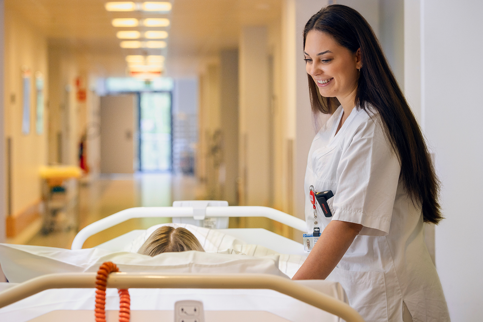 Helsearbeider ledsager pasient i seng gjennom sykehusets korridor.