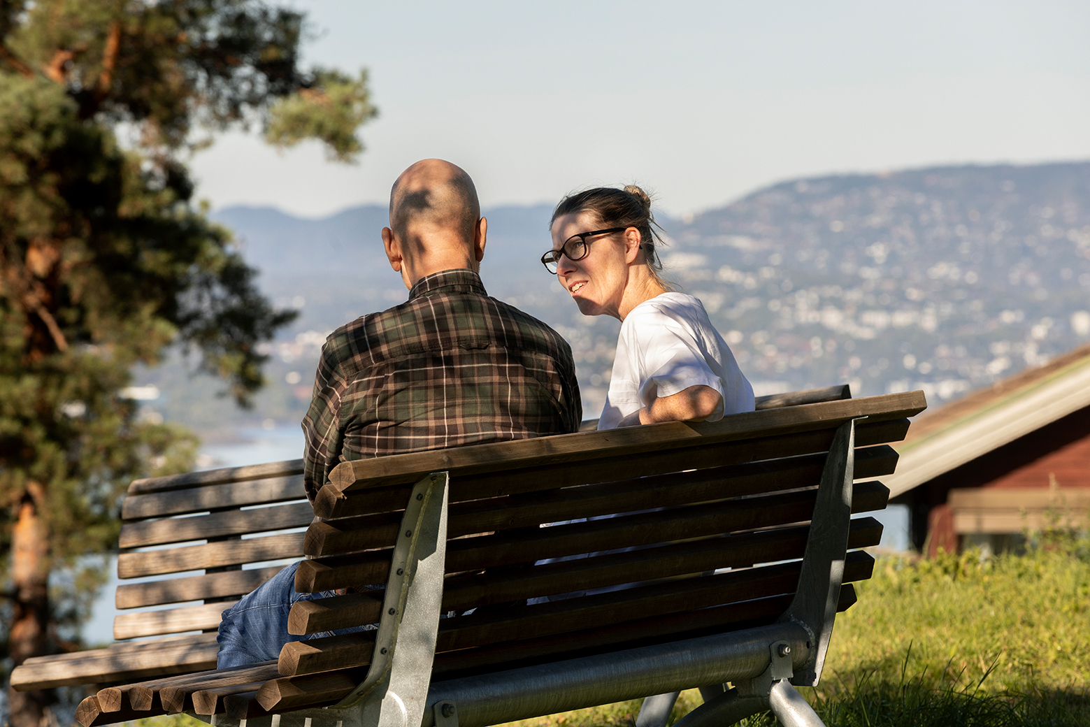 En mann og en kvinne sitter på en benk ute i naturen med utsikt over fjell og vann. De er i samtale, og solen skinner i bakgrunnen