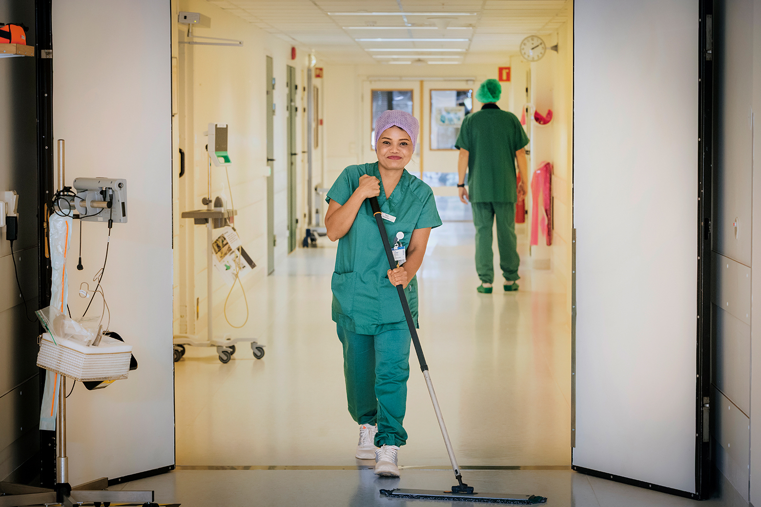 En ansatt i grønn uniform vasker gulvet i en sykehusgang for å sikre et rent og trygt miljø.