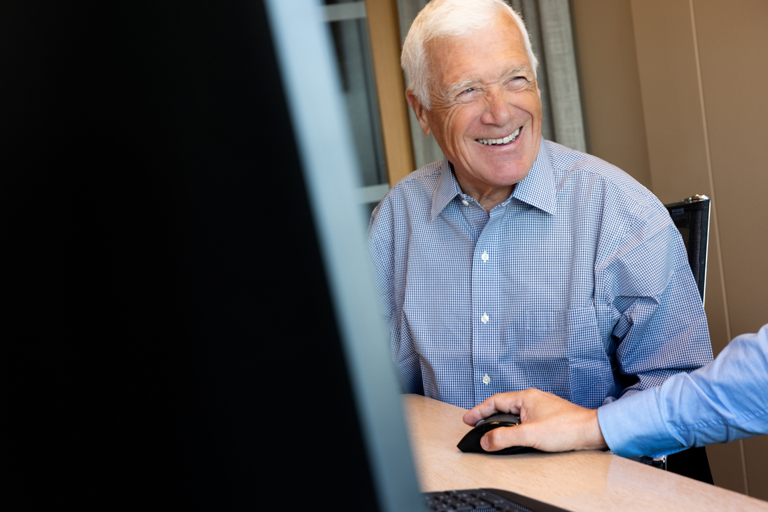 Eldre mann med hvitt hår og rutete skjorte smiler mens han sitter ved et skrivebord foran en datamaskin. En annen person ved siden av bruker datamusen