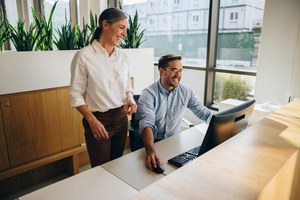 En eldre kollega mens en yngre sitter foran skjermen